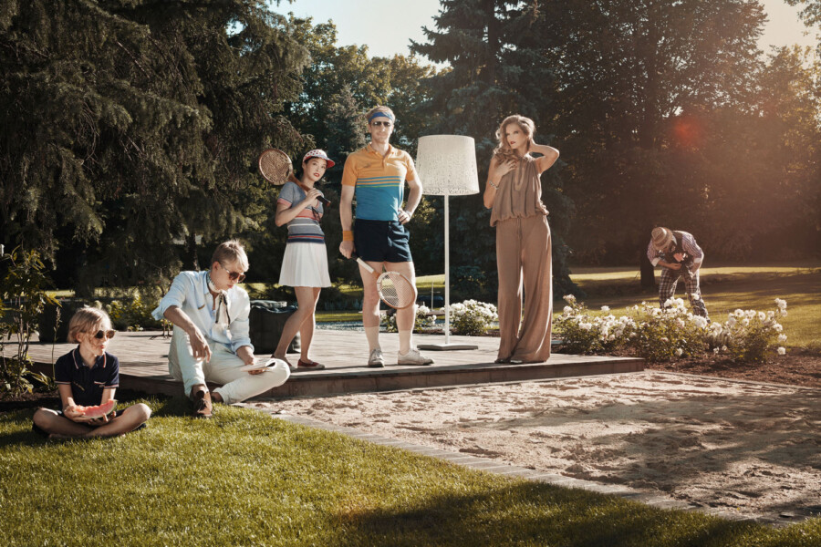 Gruppe im Park bei goldenem Licht; mehrere Personen halten Tennisschläger, eine Person im Retro-Outfit, eine weitere in braunem Overall, ein Kind isst Wassermelone.