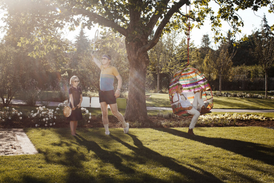 Drei Personen im sonnigen Park: links ein Kind mit Tennisschläger, Mitte eine Person im Retro-Outfit mit Schläger, rechts in einem bunten Hängesessel unter einem Baum.