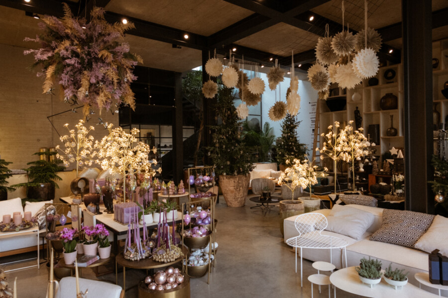 Helles Innenraumgeschäft mit weihnachtlicher Deko: beleuchtete Mini-Bäume, goldene und rosa Kugeln, weiße Möbel und hängende Papierstern-Deko.