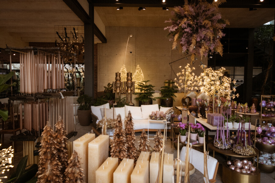 Dekorationsshop mit Kerzen, künstlichen Bäumen, Lichtern und Blütenpflanzen; weiße Couch, goldene Lichterbäume und violette Ornamente.