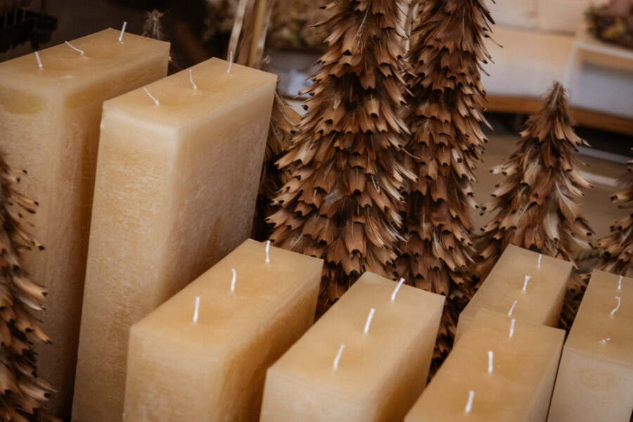 Mehrere große beigefarbene Stumpenkerzen stehen neben getrockneten braunen Tannenzweigen in einer Laden-Auslage.