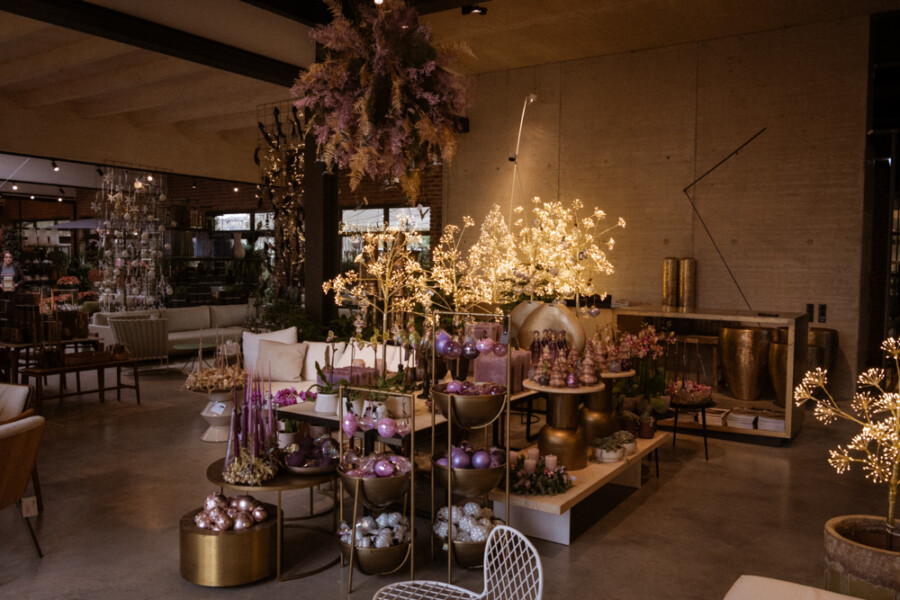 Stilvoller Innenraum mit leuchtenden Gold- und Baumdekorationen; Tische voller violetter, rosa und silberner Christbaumkugeln, moderne Möbel und warme Beleuchtung.