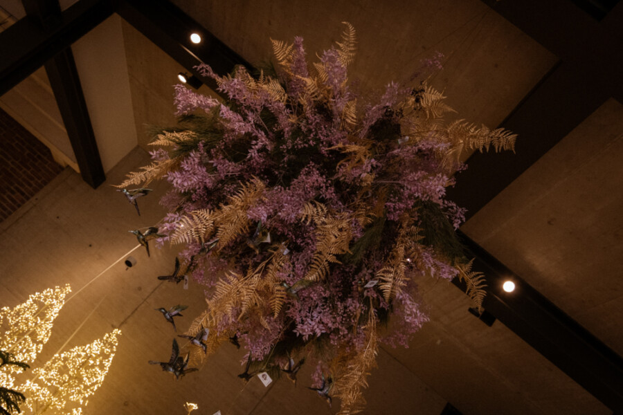 Großes hängendes Blumenarrangement mit violetten Blüten und Farnen, von der Decke eines Industriegebäudes herab, umgeben von warmen Lichterketten.