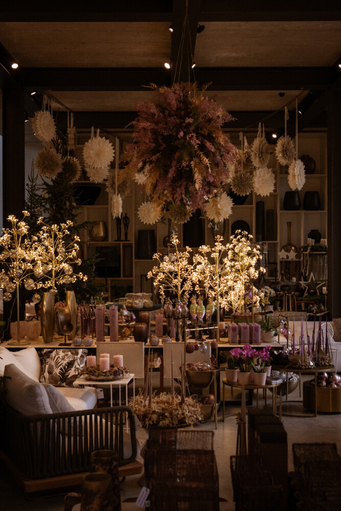 Innenraum eines Dekorationsladens: Hängebouquet aus Trockenblumen, warmes Licht, Kerzen, Vasen und Möbel auf Tischen und Regalen.
