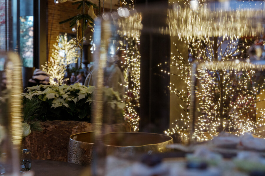Festlich beleuchteter Innenraum mit vielen Lichterketten an Zweigen, weiße Weihnachtssterne und unscharfe Glasreflexionen im Vordergrund.