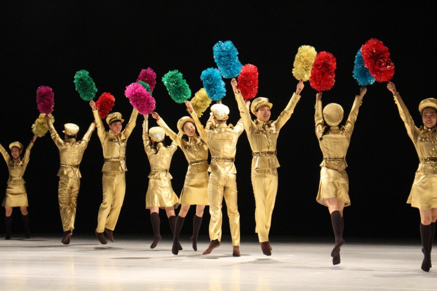 Tanzgruppe in goldenen Uniformen hebt bunte Pompons bei Bühnenauftritt vor schwarzem Hintergrund.