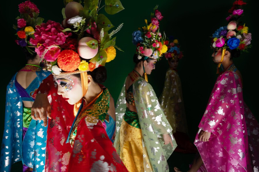 Mehrere Tänzerinnen in bunten Kimonos mit üppigen Kopfschmuck-Blumenkränzen, Hydrangea macrophylla dominierend, vor dunklem Hintergrund.