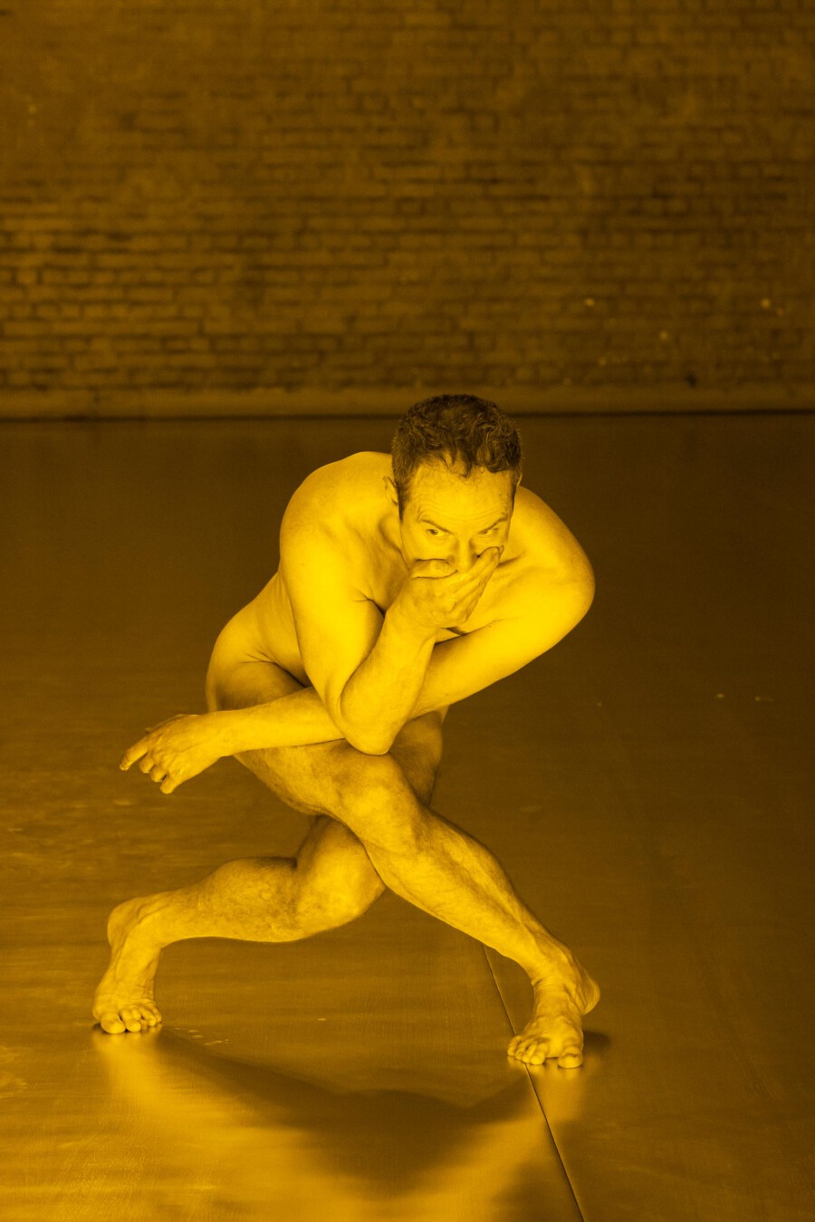 Nackte Person in hockender Pose, eine Hand am Gesicht, auf spiegelndem Boden vor Backsteinwand im Studio