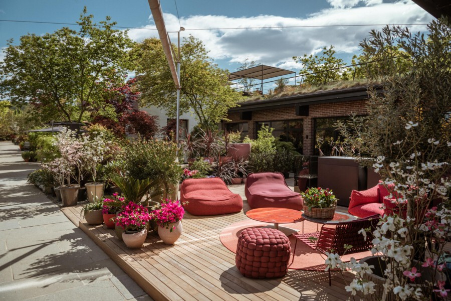 Terrasse mit Holzdielen, rote Lounge-Sessel, runder Tisch; zahlreiche Topfpflanzen, Bäume ringsum, Backsteingebäude im Hintergrund.