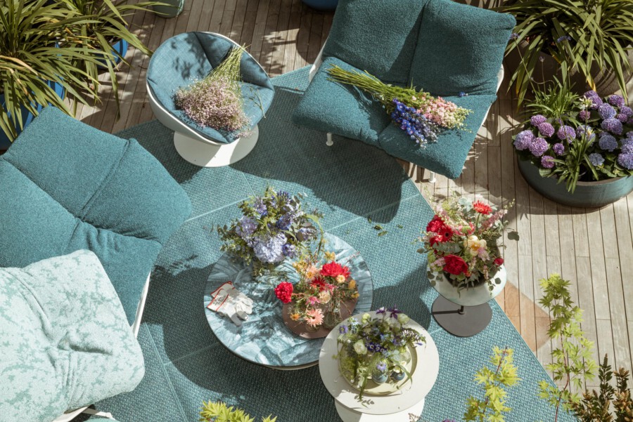 Außenterrasse mit tealfarbenen Sitzkissen; runde Tische mit frischen Blumenarrangements, darunter Allium aflatunense; Topfpflanzen umrahmen Holzdeck.